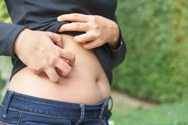 closeup of a person wearing jeans and a gray long sleeve shirt outside scratching the side of their stomach to convey the concept of an itchy bellybutton