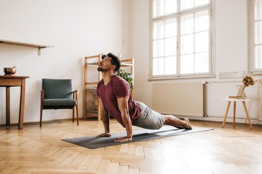 Adult in maroon T-shirt and gray shorts doing upward dog yoga pose on mat in living room.