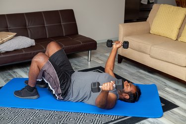 Person with short hair and sideburns holding a dumbbell in each hand, doing chest flys during a push-day workout at home, lying on a blue yoga mat next to two couches