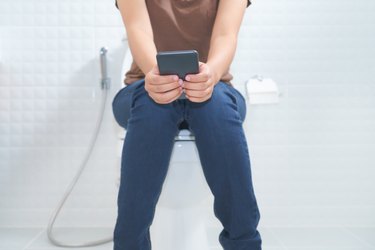 Person sitting on toilet and using smartphone, showing the concept of legs falling asleep on the toilet