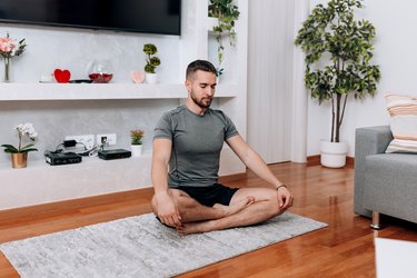 a person wearing a gray t-shirt and black shorts sits cross-legged on a gray yoga mat at home in front of plants