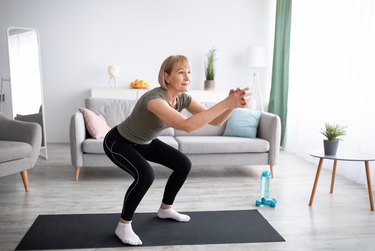 middle-aged woman doing squats on a black yoga mat in living room