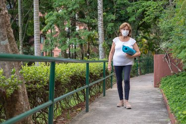 Woman walking outside to yoga class wearing face mask carrying blue mat outside