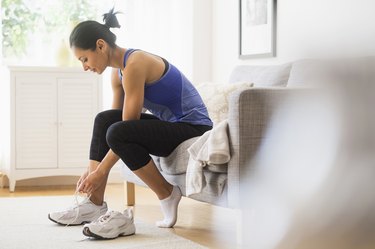 Person wearing a blue tank top and black leggings putting on exercise shoes for wide feet in living room.