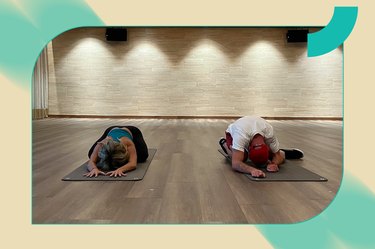 a yoga instructor and a yoga practitioner do child's pose on black yoga mats side by side in an empty studio