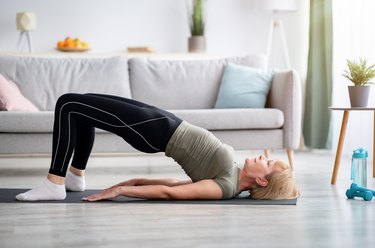 side view of a mature caucasian woman going a glute bridge exercise as part of a lower-body workout on her living room floor