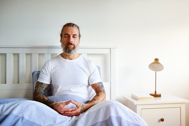an older adult wearing a white t-shirt sitting in bed under a blue sheet doing a 5 minute morning mediation to set intentions for the day next to a nightstand with a small lamp