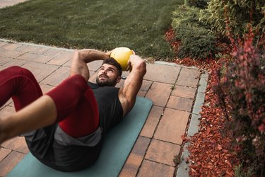 man wearing tank top and leggings doing kettlebell ab workout outside on yoga mat