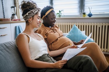 a pregnant person wearing a rust-colored dress and headband sits next to a partner on a couch at home discussing treatment plans for navigating lupus during pregnancy