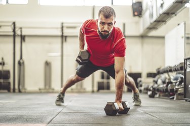 Person in a gym wearing a red shirt and black shorts doing a renegade row from a plank position while raising one dumbbell and balancing on the other