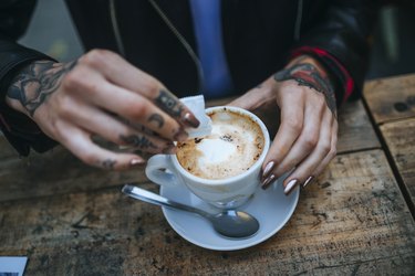 Woman's tattooed hands pouring sugar into cup of coffee, close-up