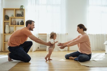 parents helping their baby daughter learn how to walk in living room
