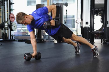 Man in a blue shirt and black shorts doing a renegade row with dumbbells during an EMOM workout in gym, exercise equipment behind him