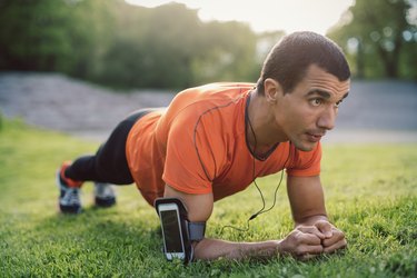 Man doing plank exercise on grassy field at park wearing smartphone armband
