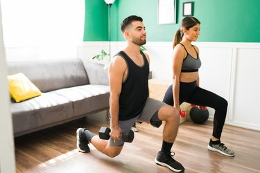 fit couple doing a dumbbell reverse lunge and working out at home together