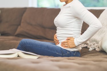 a person wearing a white long-sleeve shirt and jeans sitting on a brown couch with their hands on their pelvis because they have pelvic pain