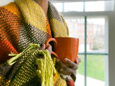 Close-up image of a person's torso wrapped in a colorful woven blanket and hands holding a bright orange mug