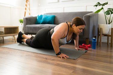 a person wearing a tank top, black leggings and a fitness tracker does a knee push-up on a gray yoga mat in their living room in front of a gray couch with a blue water bottle and red dumbbells nearby