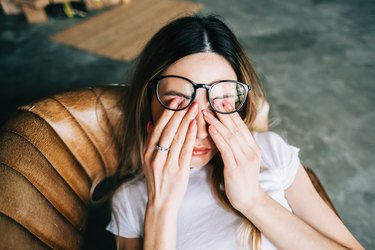 a person with long brown hair and glasses wearing a white t-shirt and rubbing their eyes because they have watery eyes